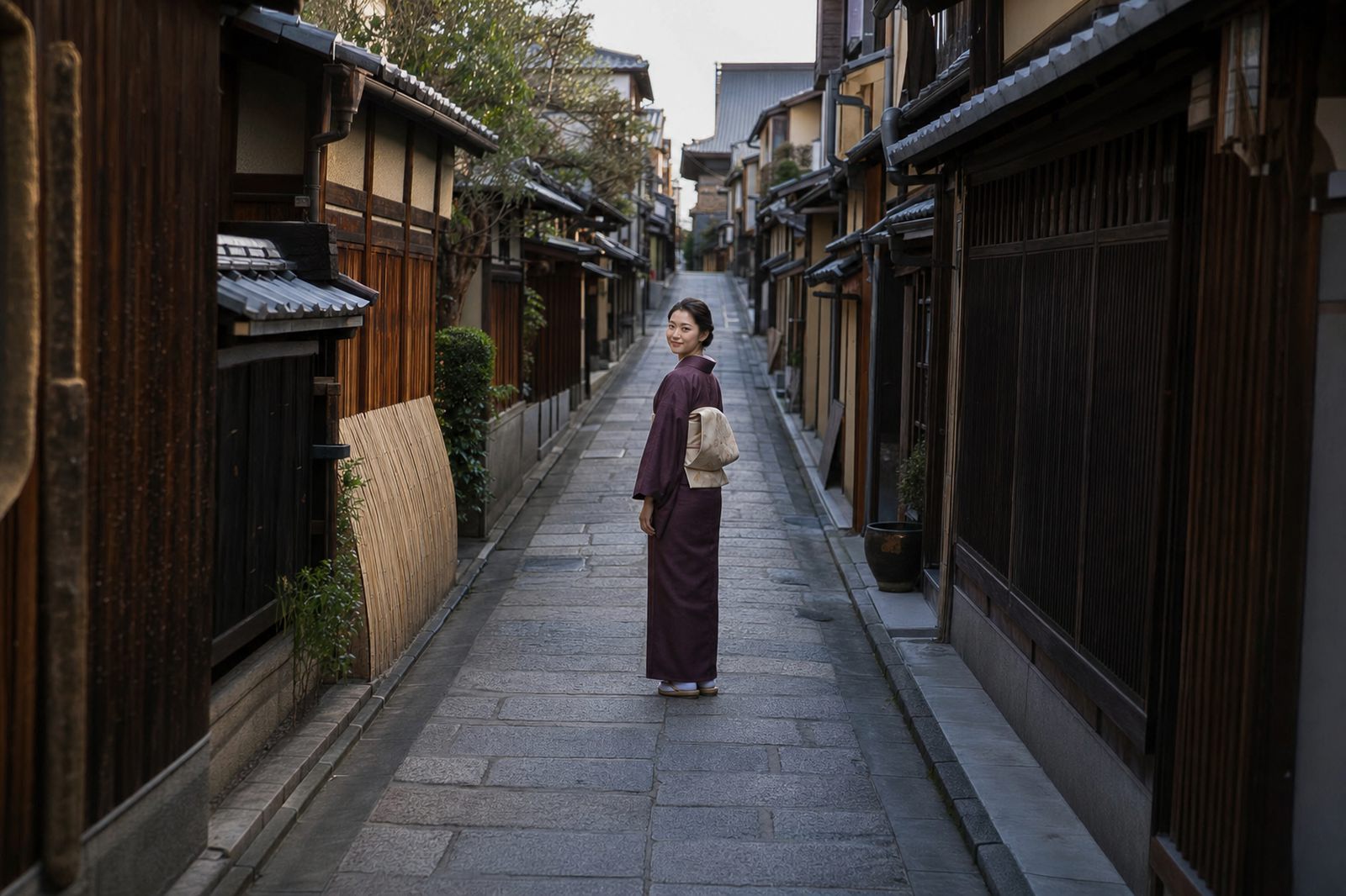 A narrow stone-paved Kyoto lane between wooden townhouses. A woman in a plum-coloured kimono has stopped in the middle of the lane and turned back over her shoulder to look at the viewer.
