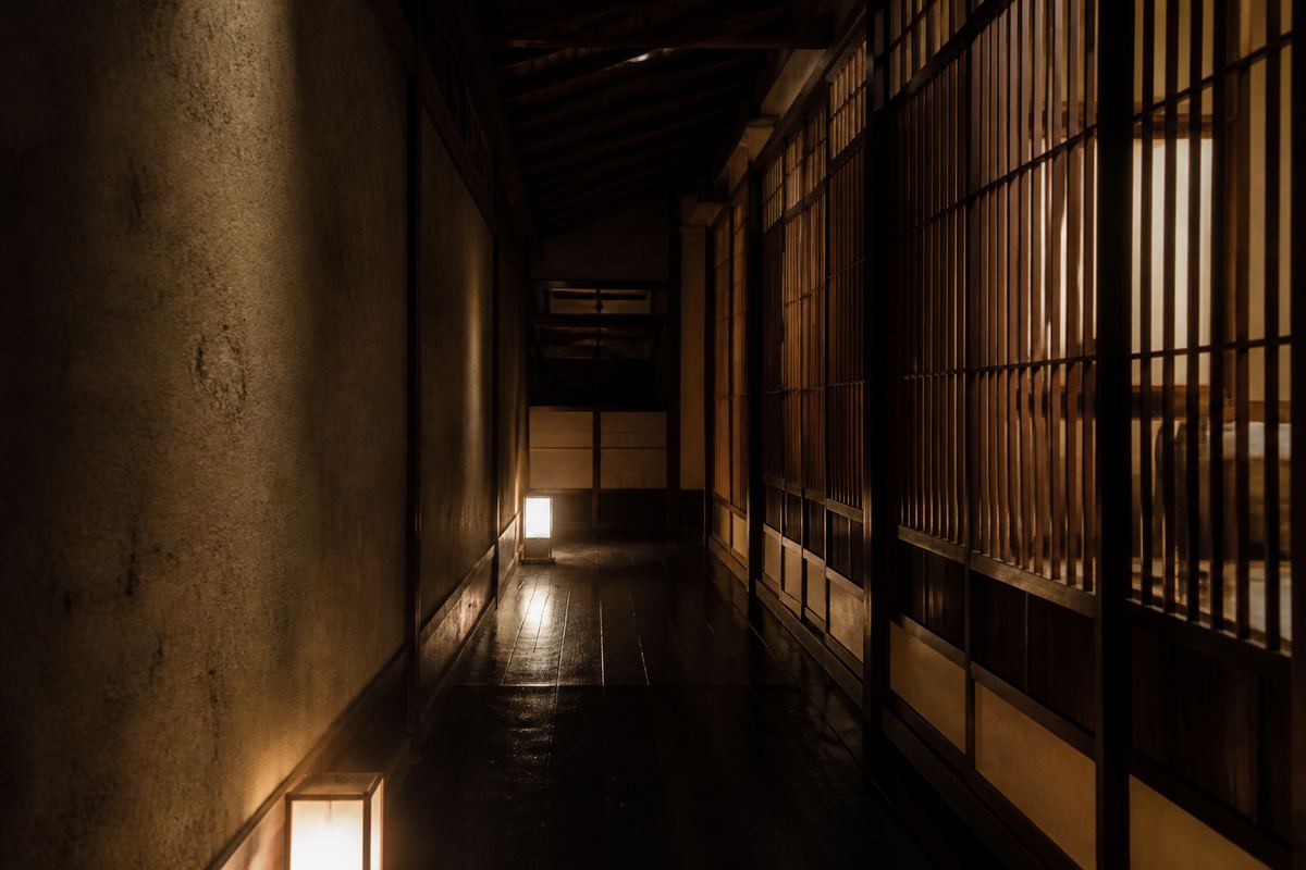 旅館のような暗い廊下。右手に格子の建具が続き、その向こうの個室からほのかに光が漏れている。