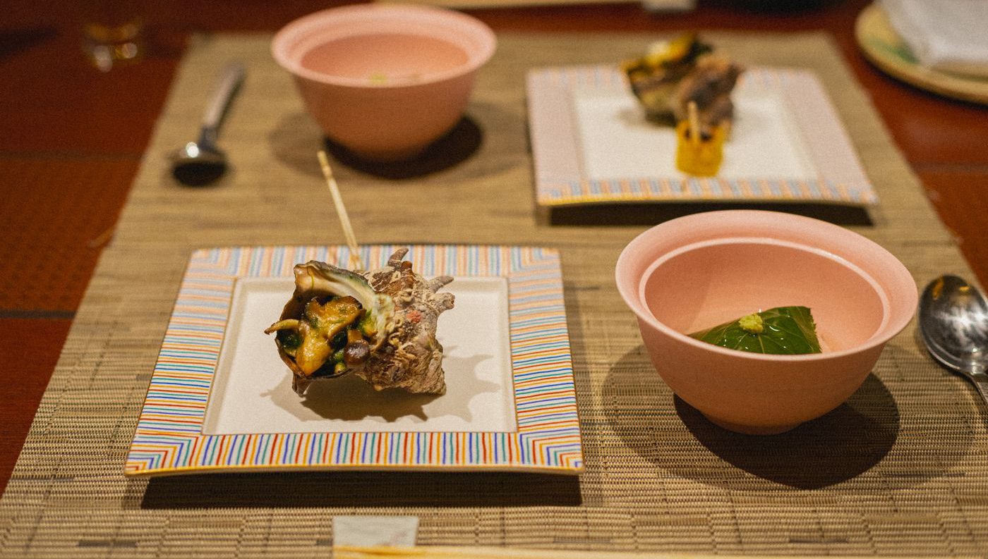 竹の敷物の上に二名分の膳。角皿に栄螺のつぼ焼き、桜色の梌に葉で包んだ蒸し物が入っている。