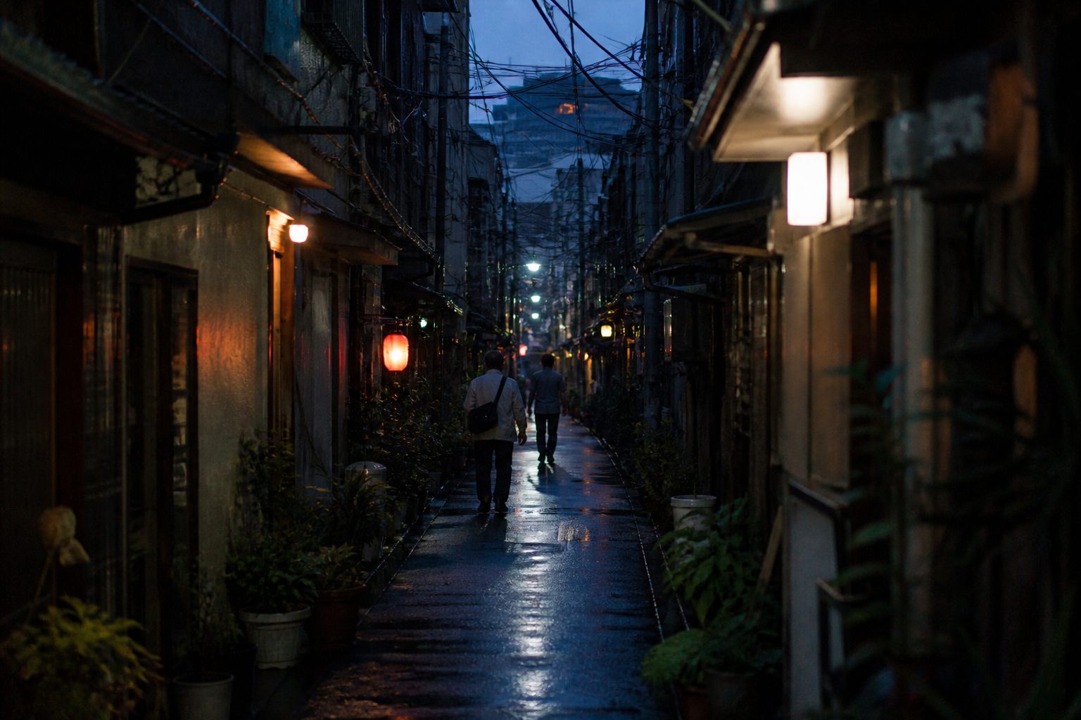 A narrow back alley at dusk. Only two people walk away in the distance; wet asphalt, overhead cables, and towers faint above the rooftops.