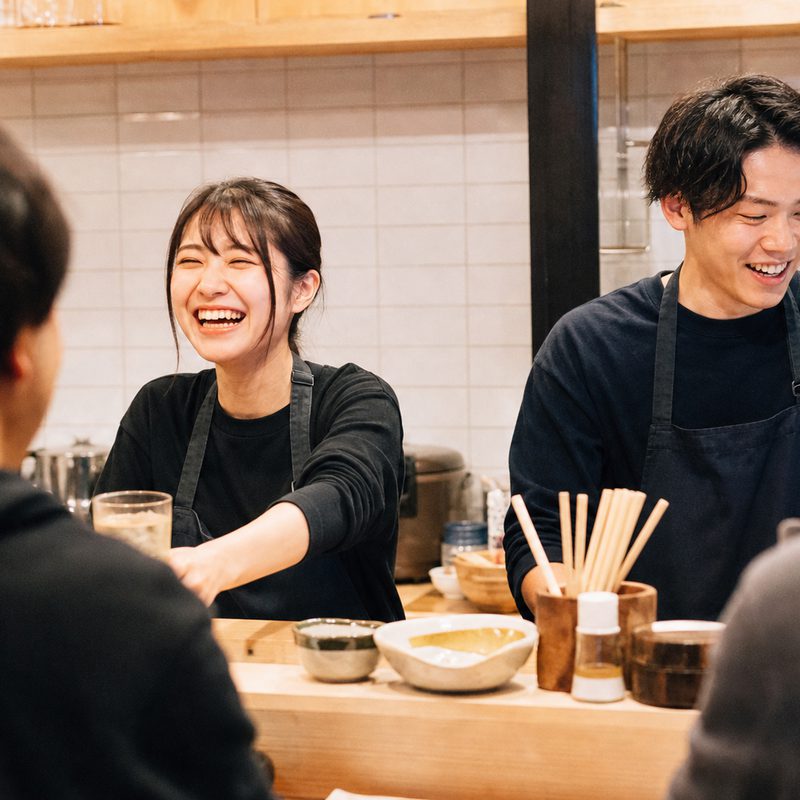 カウンターの内側で、黒いエプロンの若い女性と男性のスタッフが笑いながら客に飲み物を渡している。